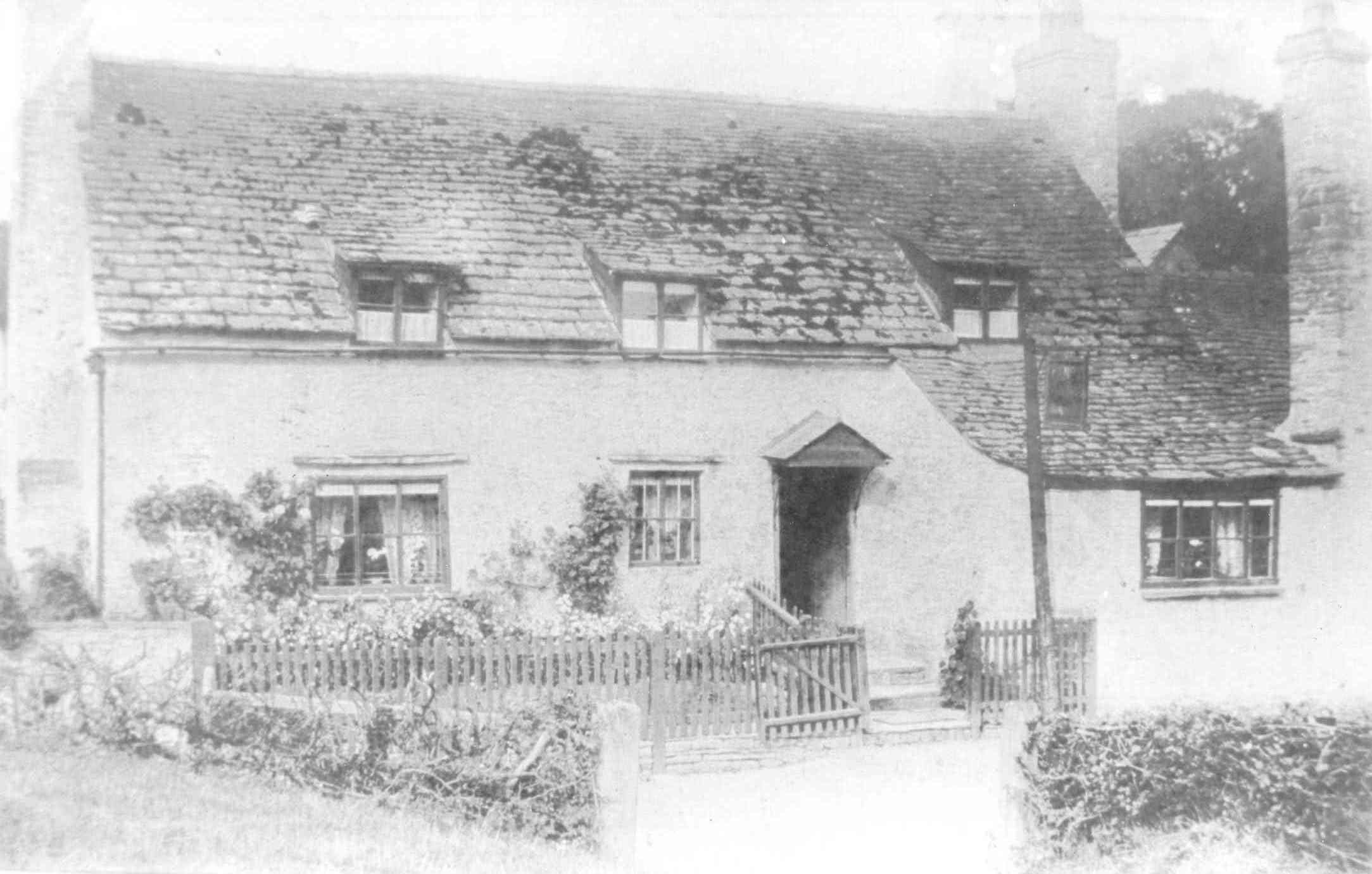 [DRAFT 2026-05-07] Edwardian monochrome photograph of a rendered cottage with a stone-tiled roof, three small dormer windows in the upper storey and a tall chimney rising at the right gable; a low picket fence with a central gate fronts the cottage, with climbing roses growing up the wall to either side of the small pent-roofed porch.