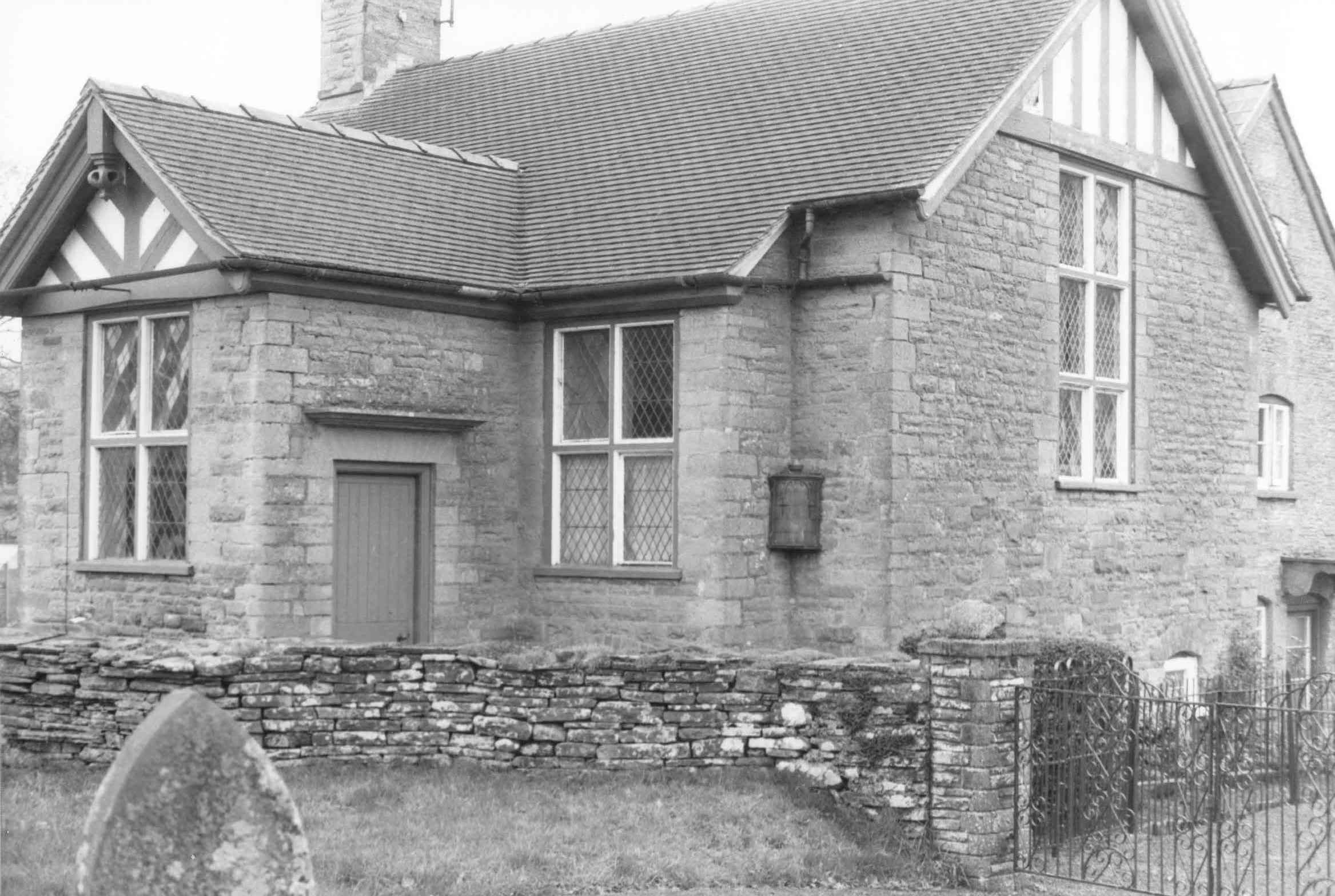 [DRAFT 2026-05-07] Stone-built single-storey reading room with a steep tiled roof, decorative half-timbered gable bargeboards and large mullioned-leaded windows on the front elevation; a low drystone churchyard wall fronts the building and a wrought-iron pedestrian gate stands to the right, with the corner of a gravestone visible at the lower left.