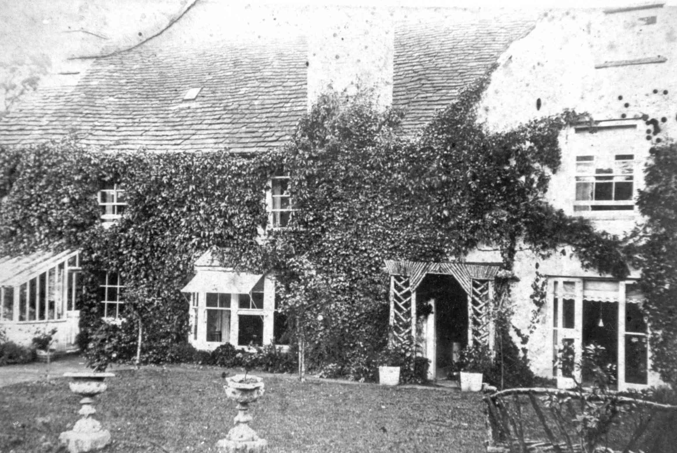 [DRAFT 2026-05-07] Aged monochrome photograph of a substantial three-bay farmhouse with a stone-tiled roof and walls almost entirely covered in ivy; a small timber-latticed porch covers the central front door, a glasshouse-like extension stands at the right gable and the foreground lawn carries two stone urns on plinths.